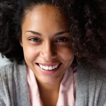 Close-up portrait of a smiling person with curly hair wearing a light pink shirt and grey sweater.