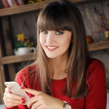 Woman with long brown hair wearing a red sweater smiles while holding a smartphone, with a wooden shelf in the background.