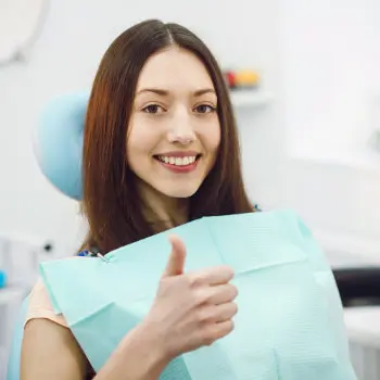 A woman sits in a dental chair wearing a bib, smiling and giving a thumbs-up.