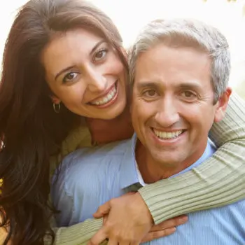 A woman with long dark hair hugs a smiling man with short gray hair from behind. Both are smiling and looking at the camera.