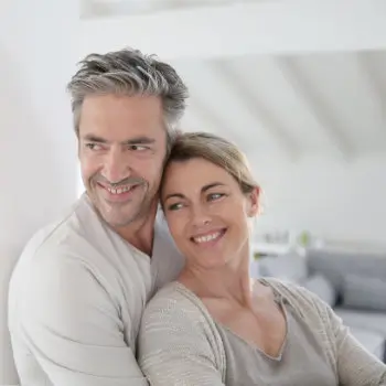 A smiling couple stands close together in a brightly lit indoor setting. The man has graying hair and the woman has blonde hair tied back. They are both casually dressed.