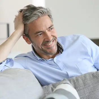 A man with graying hair and a beard, dressed in a light blue shirt, reclines on a couch while smiling and resting his head on his hand.