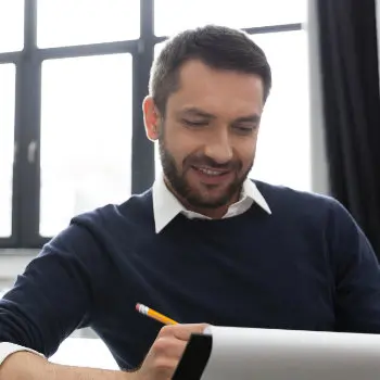 A man with short hair and a beard, wearing a dark sweater over a white shirt, smiles while writing in a notebook with a pencil. He is sitting at a desk near a window.