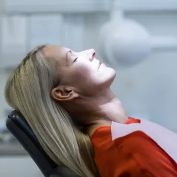 A woman with long blonde hair lies back in a dentist's chair, eyes closed, with a dental bib on, in a brightly lit dental office.