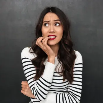 A woman with long dark hair wearing a black and white striped sweater stands against a dark background, looking anxious and biting her fingernail.