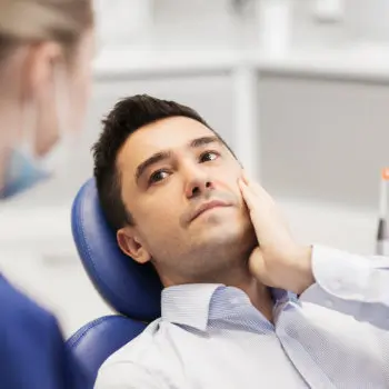 A man sits in a dental chair, touching his cheek with a concerned expression, while a dental professional stands nearby.