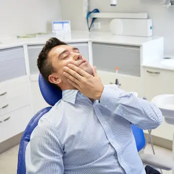 A man in a dentist's chair is touching his cheek, appearing to be in pain or discomfort. Dental tools are visible in the background.