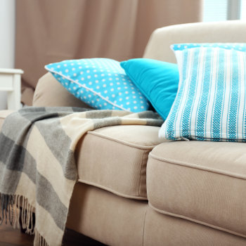 A beige couch with a gray and beige striped blanket draped over the armrest, and three blue pillows with different patterns stacked on one end.