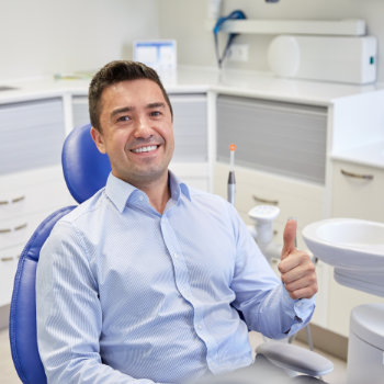 Man sitting in a dental chair smiling and giving a thumbs-up in a modern dentist office.