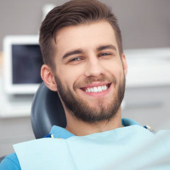 A man with a beard and mustache smiles while sitting in a dental chair, wearing a blue bib.