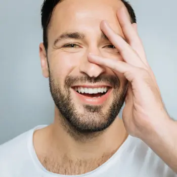 A man with short dark hair and a beard smiles while covering part of his face with his hand. He is wearing a white shirt.