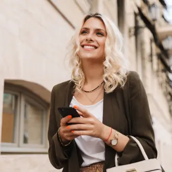 Woman with blonde hair smiling, holding a phone, standing outside near a building, wearing a dark jacket over a white top and carrying a bag.