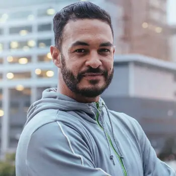 A person with short hair and a beard wearing a grey hoodie stands outdoors in front of a building, looking at the camera with a slight smile.