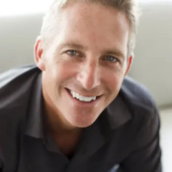 A smiling man with short, light-colored hair wearing a dark shirt looks directly at the camera.