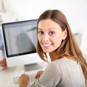 A woman with long brown hair, wearing a grey top, smiles while sitting at a desk in front of a large computer monitor.