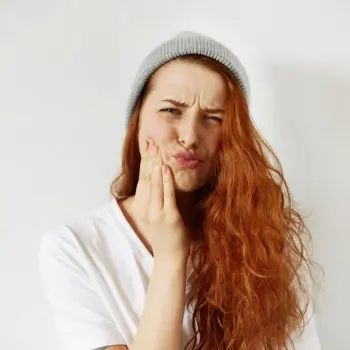 A person with long red hair and a grey beanie holds their cheek with a pained expression, as if experiencing a toothache. They are wearing a white shirt against a plain background.