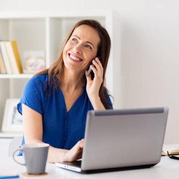 Smiling woman in a blue shirt talks on the phone while using a laptop at a desk. There is a coffee mug nearby and shelves with books and decorations in the background.