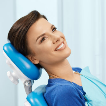 A person with short dark hair, wearing a blue top and dental bib, sits smiling in a dentist chair with a blue headrest.