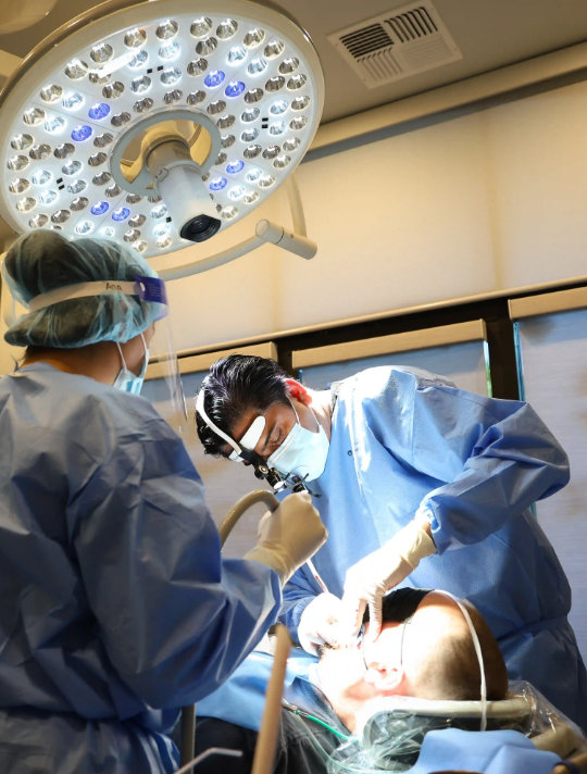 Two dental professionals in blue scrubs and masks performing a procedure on a patient under a bright overhead light.