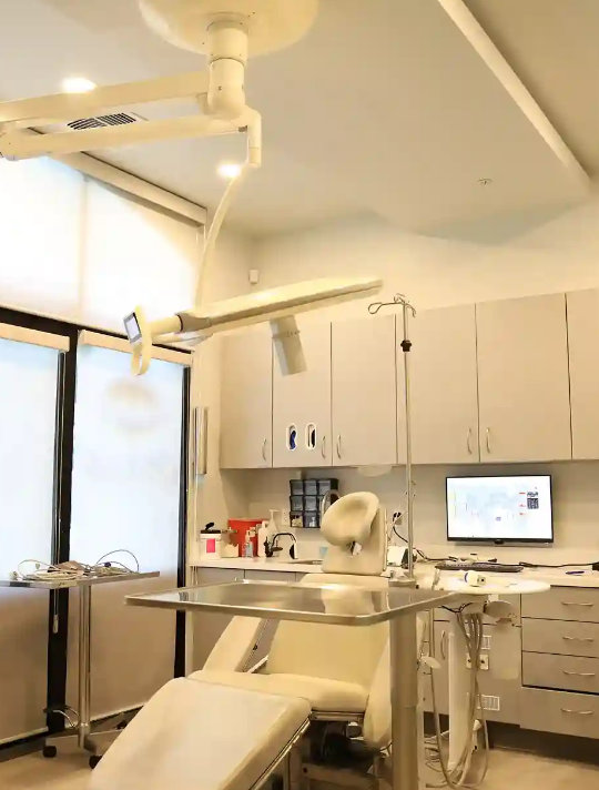 A modern dental examination room features a dental chair, overhead light, cabinetry, computer, and medical instruments on a cart.