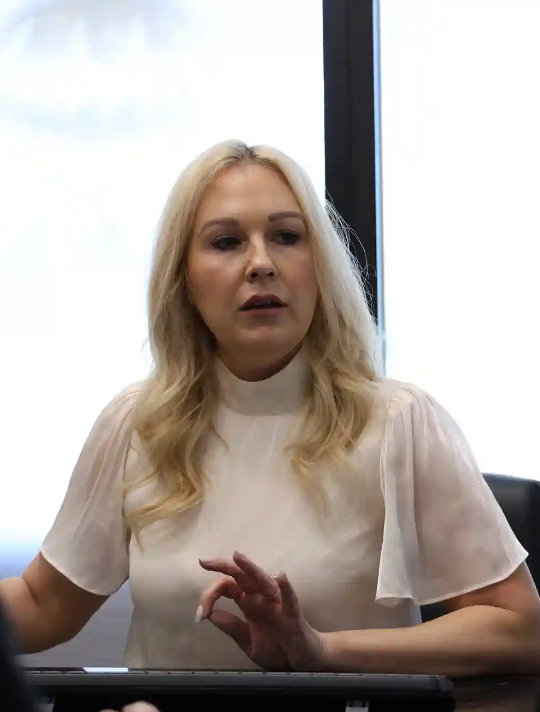 A seated woman with long blonde hair speaks, using hand gestures. She is wearing a white blouse and is positioned in front of a large window.