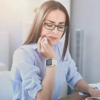 A woman with brown hair and glasses wearing a blue shirt and a white smartwatch holds her cheek, appearing to be in pain, possibly due to a toothache, while sitting indoors.