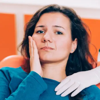 A woman seated in a dental chair, holding her cheek with a concerned expression. A gloved hand rests on her shoulder, indicating the presence of a dental professional.